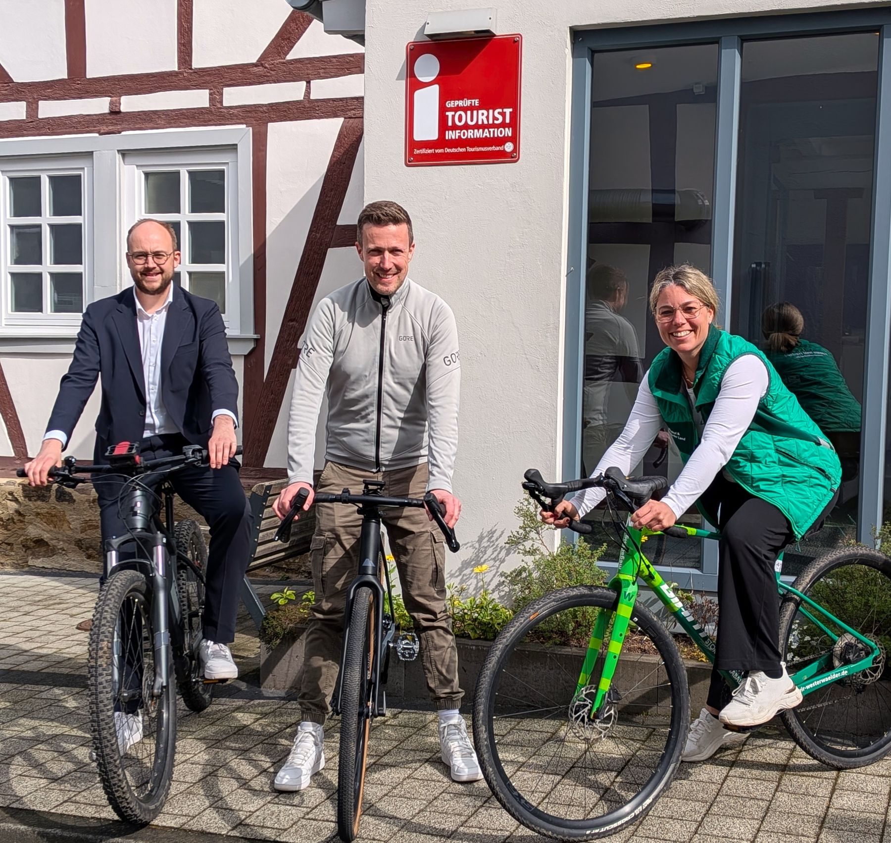 Three adults, two men and one woman, are smiling while sitting on bicycles in front of a building with a red Tourist Information sign. They are dressed in casual and business casual clothing.